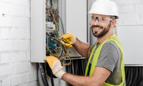 smiling handsome electrician repairing electrical box with pliers in corridor and looking at camera
